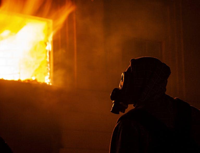 Silhouette of a person wearing a gas mask in front of a burning building at night.