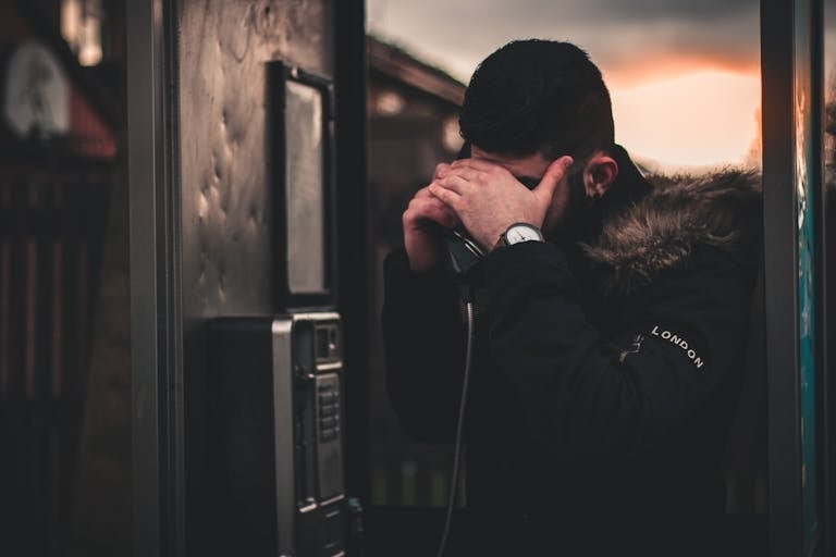 A man inside a phone booth appears worried, covering his face, during twilight.